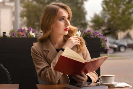 Portrait of Young modern girl, sitting at cafe outside with cup of coffee and book in the street....