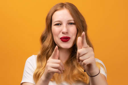 Excited girl with wavy redhead, wearing white t-shirt holding fingers in a finger gun gesture and aims at the camera. Being lucky, isolated over yellow backgroundの写真素材