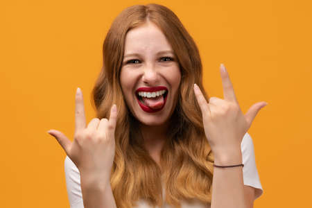 Emotive Young woman with wavy redhead makes ROCK N ROLL sign, says: I will rock this party, wearing white t-shirt, feels self confident. Human emotions conceptの写真素材