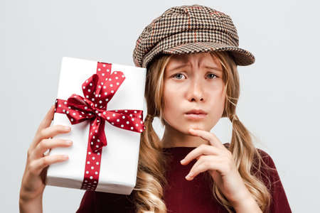 Beautiful blonde girl 10-12 years old dressed in casual sweater, holding gift box. Studio shot,...