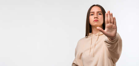 Young pretty woman with long chestnut hair, looking serious and stern, wearing casual beige hoodie, showing open palm making stop gesture. Copy space for your textの写真素材