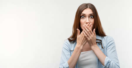 Shocked caucasian girl with long chestnut hair, dressed in casual clothes, covering mouth with hands on white background. Keep silence concept. Copy space for your textの写真素材
