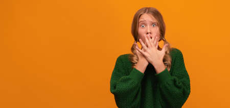 I'm afraid. Image of scared blonde girl 12-14 years old in warm green sweater covering her mouth with hands. Fright, panic attack, horror and facial expression concept. Studio shot, yellow backgroundの写真素材