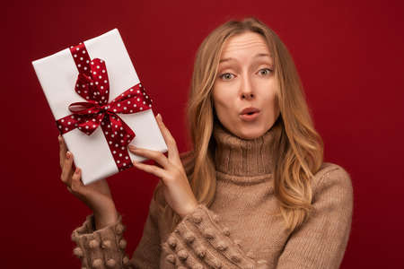 Image of charming young blonde woman in warm sweater smiling and holding gift with red ribbon....