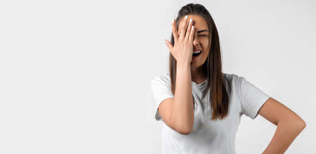 Shocked young woman with long chestnut hair dressed in casual clothes covering face with hand on...