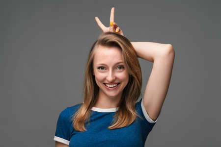 Funny young blonde female shows horns, having fun on camera. Pretty girl smiling happily, isolated on gray background. Human emotions, facial expression concept.の写真素材