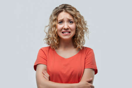Portrait of embarrassed blonde girl with curly hair in casual t shirt looking at camera. Studio...