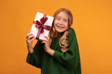 Image of charming blonde girl 12-14 years old in warm green sweater smiling and holding present box with red bow. Studio shot, yellow background. New Year Women's Day Birthday Holiday conceptの写真素材