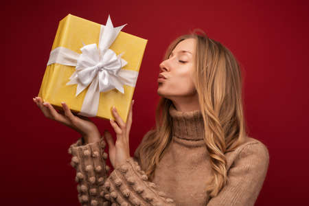 Cheerful excited young blonde woman in trendy knitted sweater smiling and holding gift. Studio shot red background. New Year Women's Day Birthday Holiday concept.の写真素材