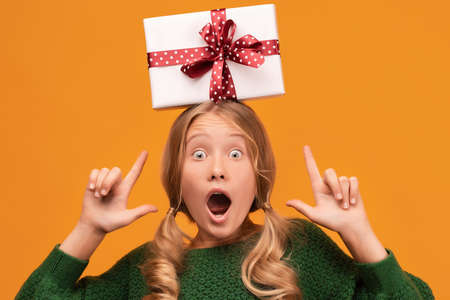 Charming blonde girl 12-14 years old in warm green sweater Hold gift box with red ribbon on her head. Studio shot, yellow background, isolated. New Year Women's Day Birthday Holiday conceptの写真素材