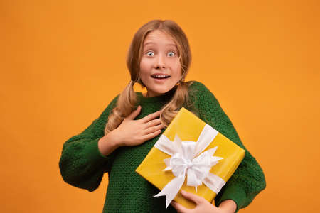 Image of charming blonde girl 12-14 years old in warm green sweater smiling and holding present box with white bow. Studio shot, yellow background. New Year Women's Day Birthday Holiday conceptの写真素材
