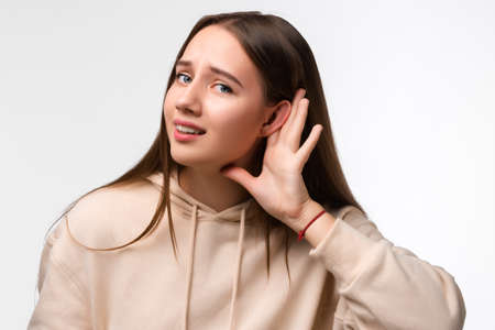 Young pretty brunette woman keeps palm of a hand close to ear as if listening to rumors isolated over white background. Human emotions, facial expression conceptの写真素材