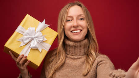 Cheerful excited young blonde woman in trendy knitted sweater smiling and holding gift. Studio shot...