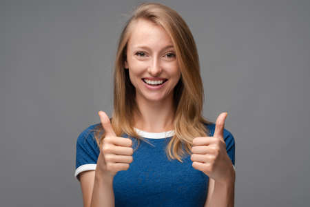 Cheerful girl with blonde straight hair looking at the camera with happy expression, showing thumbs-up with both hands, excellent job. Human emotions, facial expression concept.の写真素材
