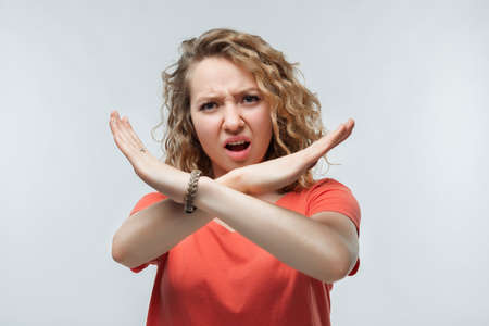 Image of serious blonde girl with curly hair in casual t shirt making X sign with crossed hands, gesturing stop, warning of danger. No way, stop doing. Studio shot, white backgroundの写真素材