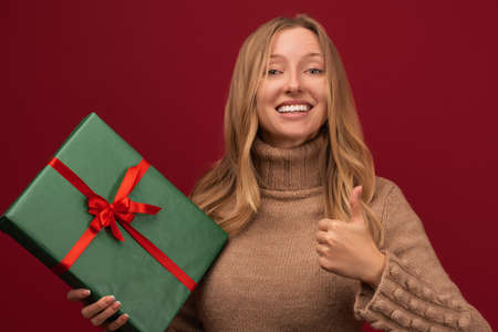 Charming young blonde woman in warm sweater shows thumb up with a present box in her hand. Studio...