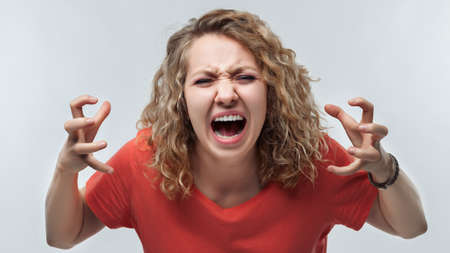 Angry blonde woman with curly hair in casual t shirt screaming and raising hands in anger. Screaming, hate, rage. Negative emotion, facial expression concept. Studio shot, white background, isolatedの写真素材