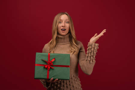 Image of charming young blonde womanin trendy knitted sweater smiling and holding gift with red...