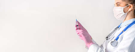 A young female doctor in a white coat, in a medical mask and pink sterile gloves hold syringe. Vaccination concept. Copy space for your textの写真素材