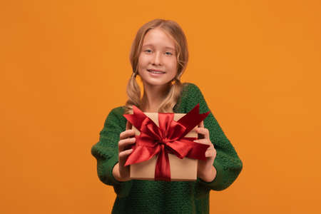Image of charming blonde girl 12-14 years old in warm green sweater smiling and holding present box with red bow. Studio shot, yellow background. New Year Women's Day Birthday Holiday conceptの写真素材