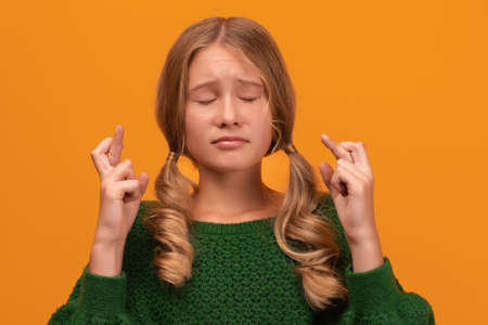 Image of pretty blonde girl 12-14 years old in warm green sweater holding fingers crossed for good luck. Human emotions, facial expression concept. Studio shot, yellow background, isolatedの写真素材