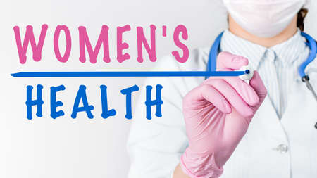 Close up female doctor in a white coat with stethoscope and pink sterile gloves writing text WOMEN'S HEALTH with marker. Medical conceptの写真素材