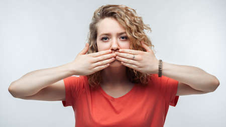 Image of blonde girl with curly hair in casual t shirt shutting mouth palms of the hand, promises to keep secret. Silence and secret concept. Studio shot, white background, isolatedの写真素材