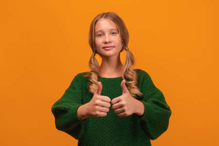 Image of charming blonde girl 12-14 years old in warm green sweater smiling and showing thumbs up....