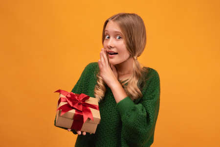 Image of charming blonde girl 12-14 years old in warm green sweater holding present box with red bow. Studio shot, yellow background, isolated. New Year Women's Day birthday holiday conceptの写真素材