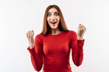 Image of young happy woman with long chestnut hair, wearing in casual red sweater smiling and making winner gesture, celebrating amazing news. Studio shot, white background, isolatedの写真素材