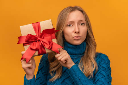 Portrait of a charming young woman in knitted blue sweater holding gift decorated with ribbon. Studio shot, yellow background. New Year, Women's Day, Birthday, Holiday conceptの写真素材