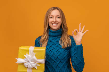 Happy young woman in knitted blue sweater shows OK gesture with a present box in her hand. Studio...