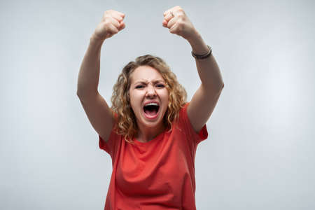 Image of young happy woman with curly hairstyle, dressed in casual t-shirt smiling and making winner gesture, celebrating amazing news. Studio shot, white background, isolatedの写真素材