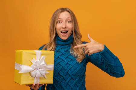 Portrait of a happy young woman in knitted blue sweater holding gift decorated with ribbon. Studio shot, yellow background. New Year, Women's Day, Birthday, Holiday conceptの写真素材