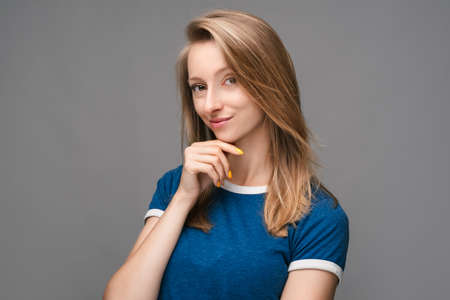 Thoughtful young female with blonde straight hair and in a blue t shirt, looking confident at the camera with hand raised on chin, isolated over gray background. Concept of thinkingの写真素材