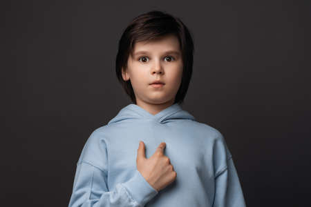 Photo of puzzled boy 10-12 years old, dressed in casual clothes pointing finger at herself, has dissatisfied expression. Studio shot, gray background. Human emotions, facial expression conceptの写真素材