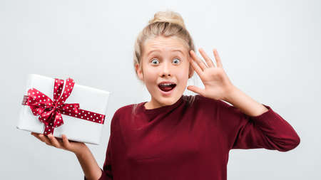 Charming blonde girl 12-15 years old with gift decorated with ribbon posing with surprised face expression. Studio shot white background, isolated. New Year, Women's Day, Birthday, Holiday conceptの写真素材