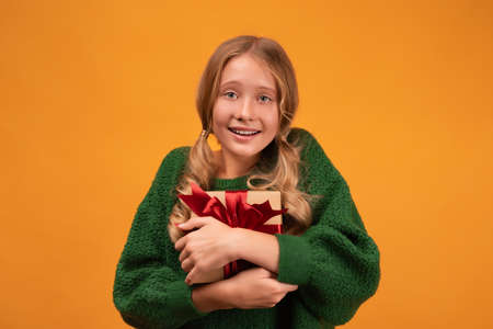 Image of charming blonde girl 12-14 years old in warm green sweater smiling and holding present box...
