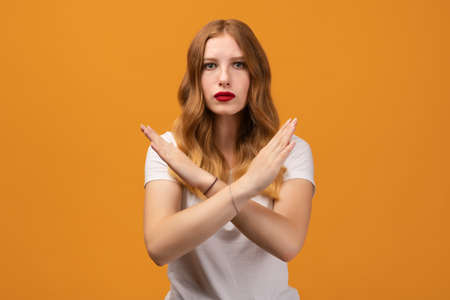 Image of serious woman 20s with wavy redhead, making X sign with crossed hands, gesturing stop, warning of danger. No way, stop doing. Wearing white t-shirtの写真素材