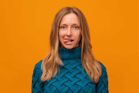 Portrait of cute young woman in knitted blue sweater smiling and sticking out her tongue at camera....