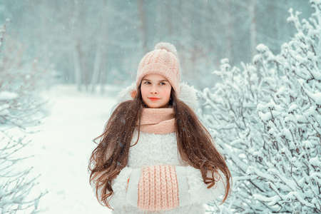 Beautiful little girl 10-12 years old in knitted hat on winter forest background. Warm white winter...