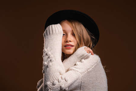 Charming blonde teen girl in black hat posing in studio on brown background. Concept of beauty and eleganceの写真素材