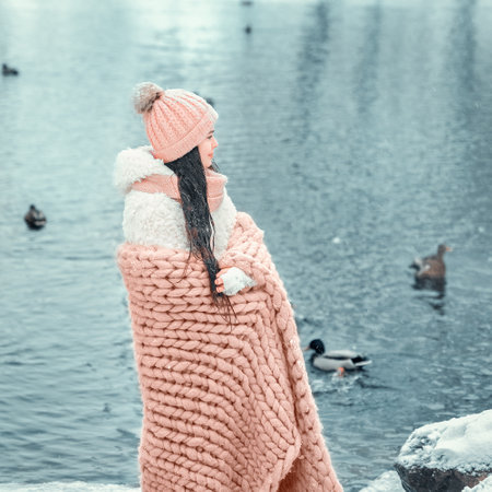 Little girl 10-12 years old wrapped in warm blanket standing and looking at lake. Cold morning by the lake. Image with selective focus, toning and noiseの写真素材