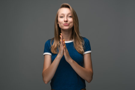 I am burning from anticipation. Good-looking woman with blonde straight hair wears blue t shirt, clasping hands near face and being impatient and excited about something over gray wallの写真素材
