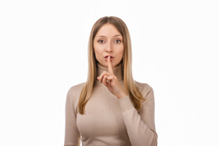 Portrait of young blond woman keeping finger at her mouth, saying Shh, Hush, Tsss. Silence and secret concept. Studio shot, white backgroundの写真素材