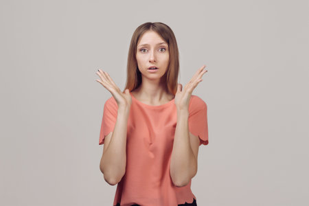 Worried and concerned blond girl in casual clothes is looking with surprised expression, clueless how act, feel embarrassed and worried. Studio shot, gray backgroundの写真素材