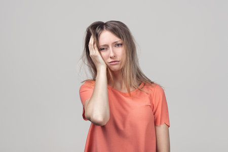 Tiredness concept. Displeased sleepy girl feels fatigue, covers face with hand, wakes up very early, suffers from headache. Studio shot, gray backgroundの写真素材