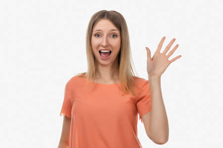 Friendly blond girl in casual clothes is saying hello, smiling joyfully and friendly, waving her hand, showing five fingers. Studio shot, white backgroundの写真素材