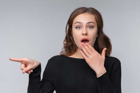Portrait of young brunette woman, pointing left and inviting people look promo, standing against gray background. Lovely brunette girl looks surprisedの写真素材