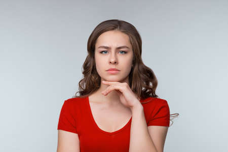 Hmm, interesting. Thoughtful young brunette woman, looking at camera and thinking about something, making assumption, pondering. Studio shot, gray background. Human emotions, facial expression conceptの写真素材